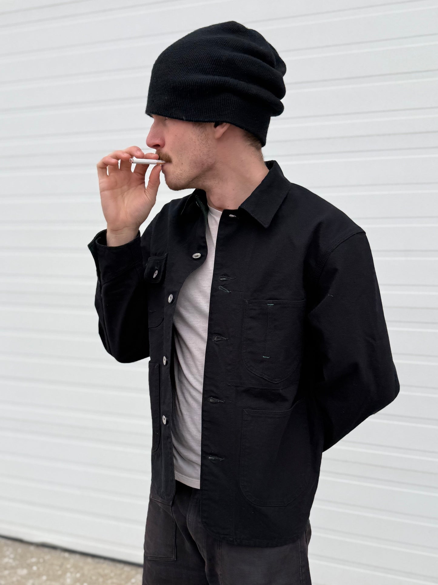 Seb wearing a Keymaster black chorecoat, beanie, and pants smoking against a white wall.