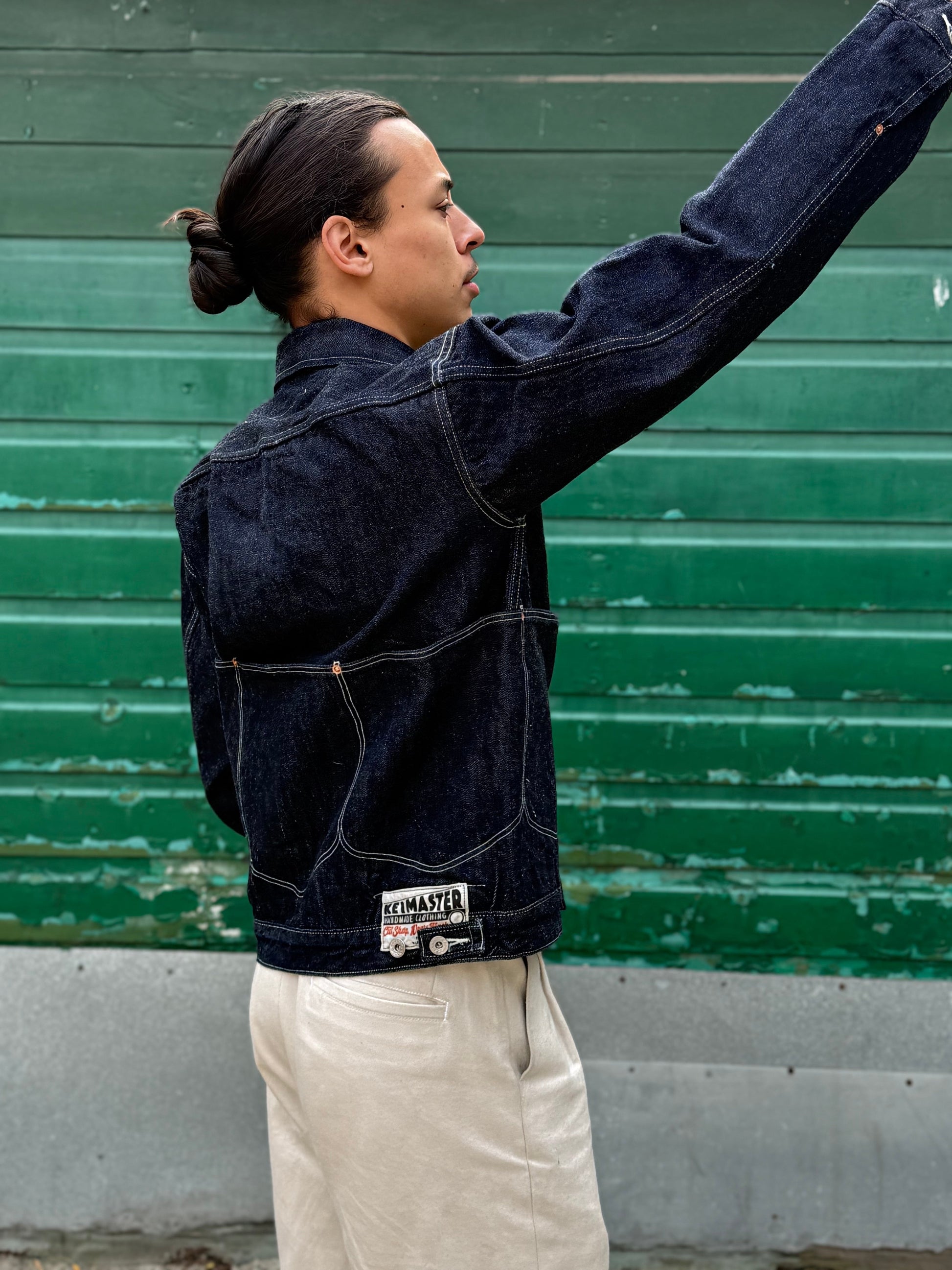 Elio wearing a dark denim jacket with a visible brand label against a green wall.