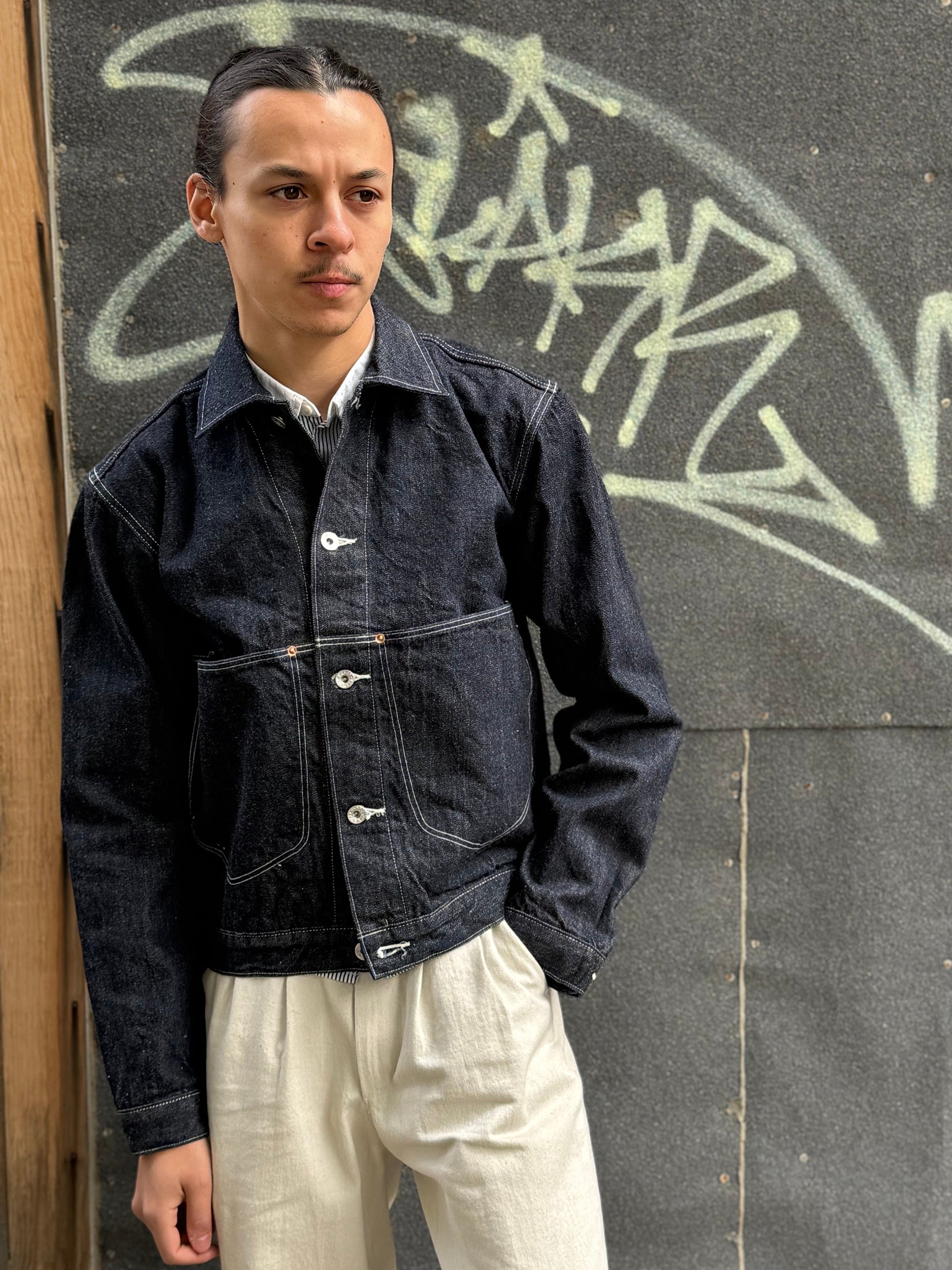 Elio wearing a dark denim jacket and light-colored pants standing against a wall with graffiti.
