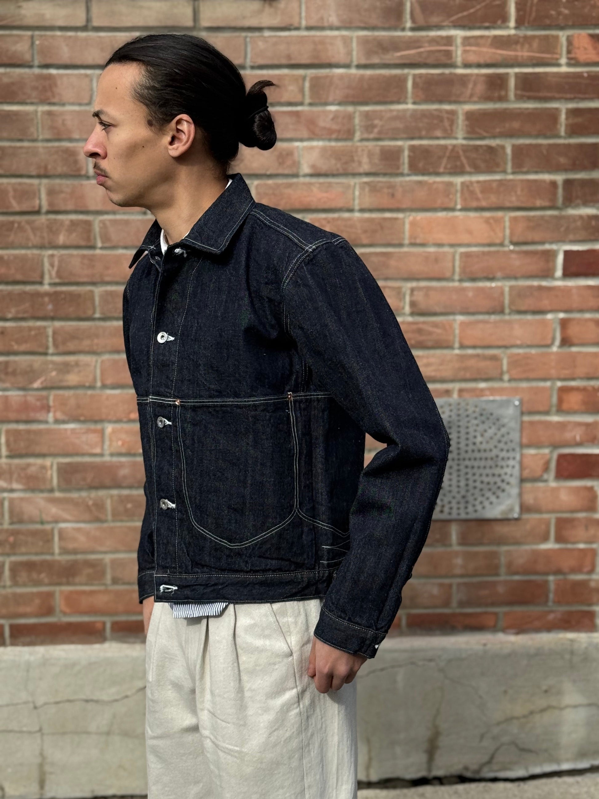 Elio wearing a dark denim jacket in size 42 and light-colored pants against a brick wall.