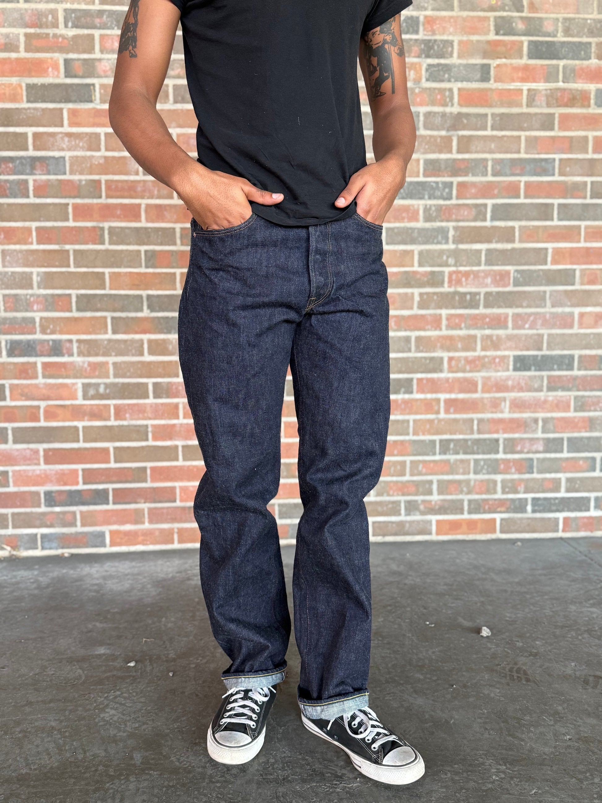 Person wearing dark jeans and black shoes standing against a brick wall.